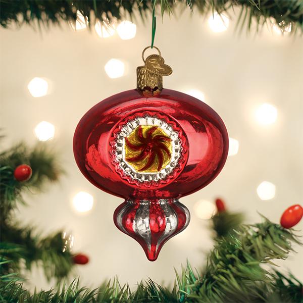 A shiny red Christmas ornament with gold and yellow accents hangs on a green Christmas tree branch, with blurred lights glowing softly in the background.