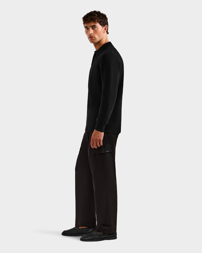 A man with short brown hair stands sideways, looking at the camera, wearing a black Rudsak RICHARD - 6524529 long sleeve polo, dark pants, and black shoes against a plain light gray background.