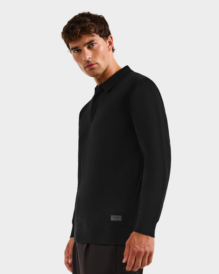 A young man with short brown hair models the Rudsak RICHARD - 6524529 Black men's knit polo sweater and dark pants against a plain light background, looking slightly to the side—an ideal addition to any modern wardrobe.