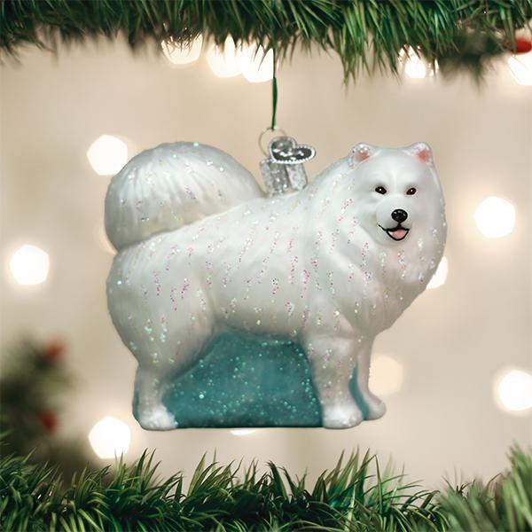 A Christmas tree ornament shaped like a fluffy white dog, possibly a Samoyed, with glitter accents hangs among green tree branches, with soft white lights blurred in the background.