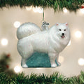 A Christmas tree ornament shaped like a fluffy white dog, possibly a Samoyed, with glitter accents hangs among green tree branches, with soft white lights blurred in the background.