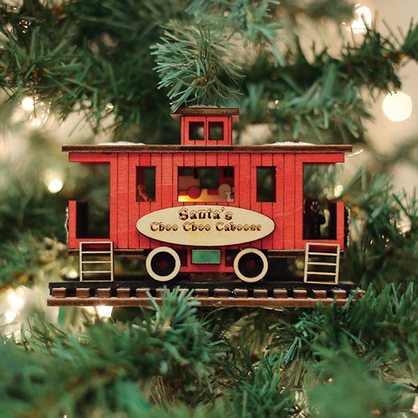 A red wooden train caboose ornament labeled Santas Choo Choo Caboose hangs on a Christmas tree, surrounded by green branches and twinkling white lights.