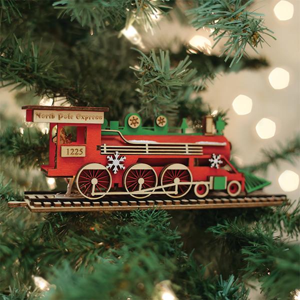 A wooden â€œNorth Pole Expressâ€_x009d_ train ornament, painted red and green with snowflake decorations, hangs on a Christmas tree with blurred white lights in the background.
