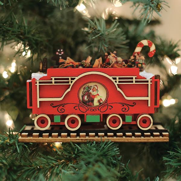 A red and green wooden train ornament with a Santa Claus image and toys on top hangs on a Christmas tree with twinkling lights in the background.