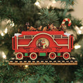 A red and green wooden train ornament with a Santa Claus image and toys on top hangs on a Christmas tree with twinkling lights in the background.