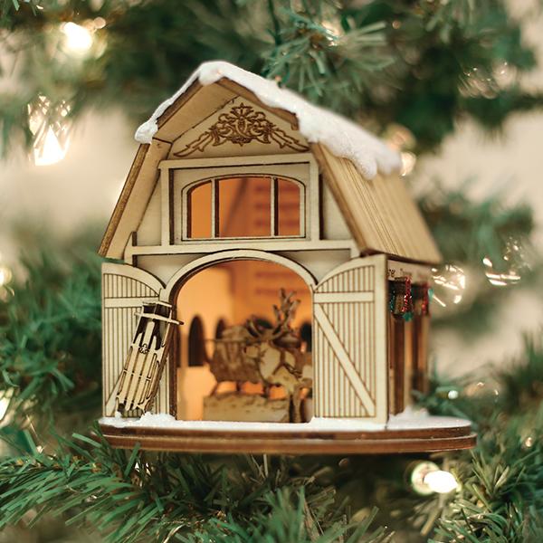 A wooden Christmas ornament shaped like a barn, decorated with a sled and greenery, hangs on a Christmas tree. Inside, a reindeer figure is visible, with warm glowing lights creating a cozy atmosphere.