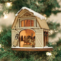 A wooden Christmas ornament shaped like a barn, decorated with a sled and greenery, hangs on a Christmas tree. Inside, a reindeer figure is visible, with warm glowing lights creating a cozy atmosphere.