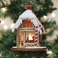 A Christmas tree ornament shaped like a snow-covered Santaâ€™s Workshop with glowing windows, a red-and-white candy cane, and a gingerbread figure, hanging on a tree with twinkling lights in the background.