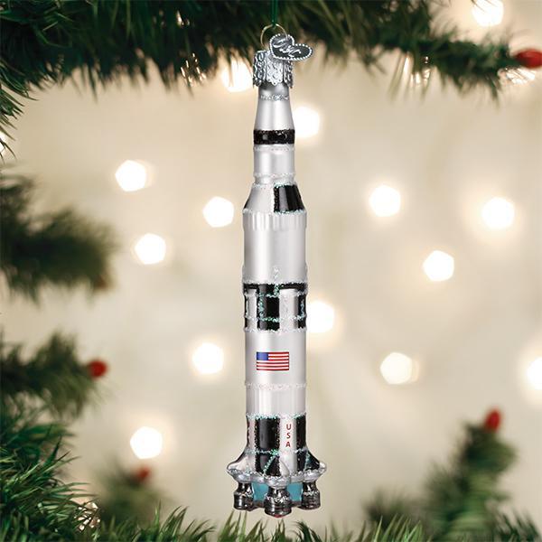A Christmas tree ornament shaped like a silver and white rocket with a USA flag, hanging on a green tree branch, with blurred holiday lights in the background.