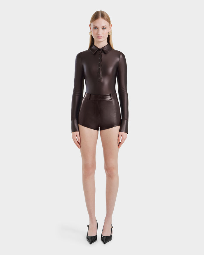 A woman faces forward against a plain white background, wearing the Rudsak SHELLY - 8524537 Espresso stretch fabric bodysuit with long sleeves and a collar, paired with matching high-waisted shorts and black pointed high heels.