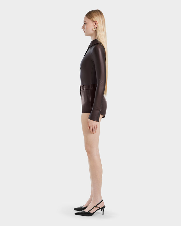 A woman with long straight blonde hair stands in profile, wearing the Rudsak SHELLY - 8524537 Espresso womens bodysuit with long sleeves and a belt, paired with black heeled slingback shoes against a plain white background.