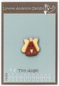 A hand painted wooden button with a design resembling a tiny angel, placed on a grey background with a fence-like pattern.