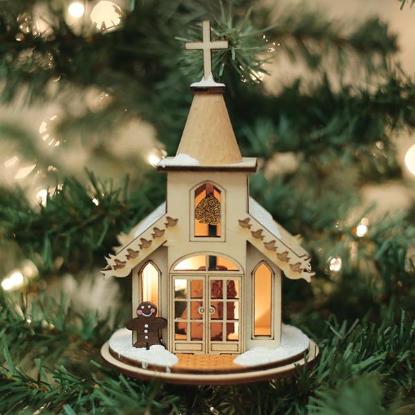 A small, illuminated wooden church ornament with a cross on top, snow accents, and a gingerbread figure stands in front. The ornament hangs on a Christmas tree with blurred lights in the background.