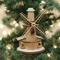 A wooden windmill ornament decorated with a small wreath hangs on a Christmas tree, surrounded by green branches and soft, blurred holiday lights in the background.