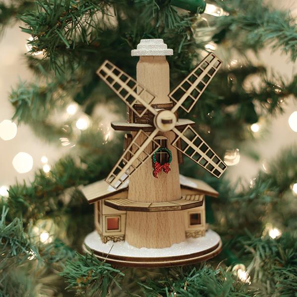 A wooden windmill ornament decorated with a small wreath hangs on a Christmas tree, surrounded by green branches and soft, blurred holiday lights in the background.